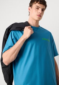 Τυρκουάζ βαμβακερό T-shirt με άνετη εφαρμογή και κοντά μανίκια, διαθέτει πράσινο λογότυπο στο στήθος. Μαύρο μπουφάν κρεμασμένο στον ώμο του μοντέλου.