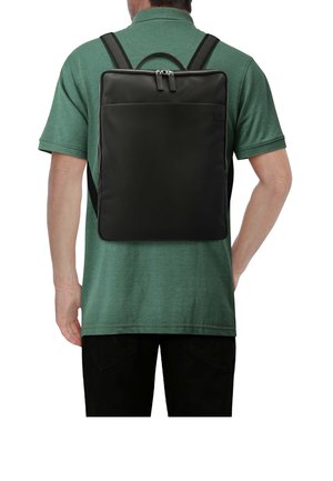 Mann trägt grünes Polo und schwarze Hose mit einem eleganten schwarzen rechteckigen Rucksack, der einen oberen Reißverschluss und eine Fronttasche hat, von hinten gesehen.