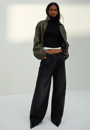 Žena stojí s rukami v vreckách, má na sebe olivovo zelenú koženú bundu, čierny krátky rolák, široké čierne nohavice a čierne špicaté topánky.