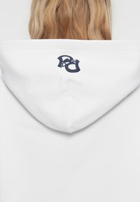 Hoodie bianco con un interno grigio testurizzato e logo blu navy sulla parte posteriore del cappuccio. Design semplice senza ulteriori motivi o dettagli.