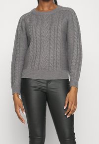 Pull en maille grise avec un motif torsadé, col rond, poignets et ourlet côtelés. Porté avec un pantalon noir en similicuir.