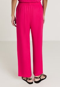 Frau in leuchtend pinkfarbener, weit geschnittener Hose und schwarzen Sandalen mit Riemen, steht gegen einen schlichten hellen Hintergrund, Rückansicht.