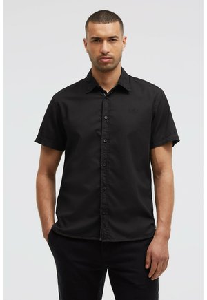 Mann mit kurzem Haar, trägt ein kurzärmeliges schwarzes Hemd mit Knöpfen, steht mit den Händen in den Taschen, neutraler Gesichtsausdruck, hellgrauer Hintergrund.