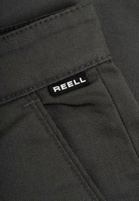 Dark gray fabric with a ribbed texture and visible stitching. Features a black label with white text reading "REELL."