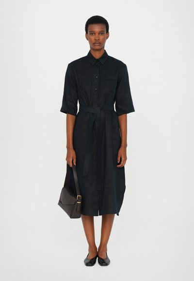 Vestido preto de camisa com botões, um cinto combinando e mangas curtas. Combinado com uma bolsa escura e sapatos planos pretos. Design simples.