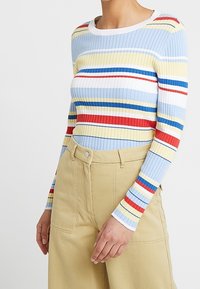 Ribbed pitkähihaine paita, jossa on vaakasuorat raidat sinisen, keltaisen, punaisen ja valkoisen yhdistelmässä. Yhdistetty korkeavyötäröisten beigejen housujen kanssa.