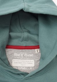 Innenseite eines türkisfarbenen Baumwollhoodies mit einem grauen Etikett, auf dem der Markenname, die Größenangaben und Pflegehinweise stehen. Roter Akzent entlang des Halsausschnitts.