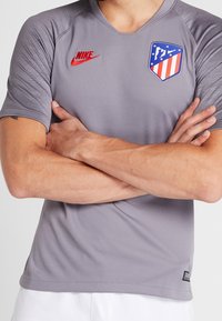 Šedé sportovní tričko Nike s véčkovým výstřihem, krátkými rukávy, texturovanými akcenty na ramenou a barevným týmovým logem. Červené logo Nike na hrudi.