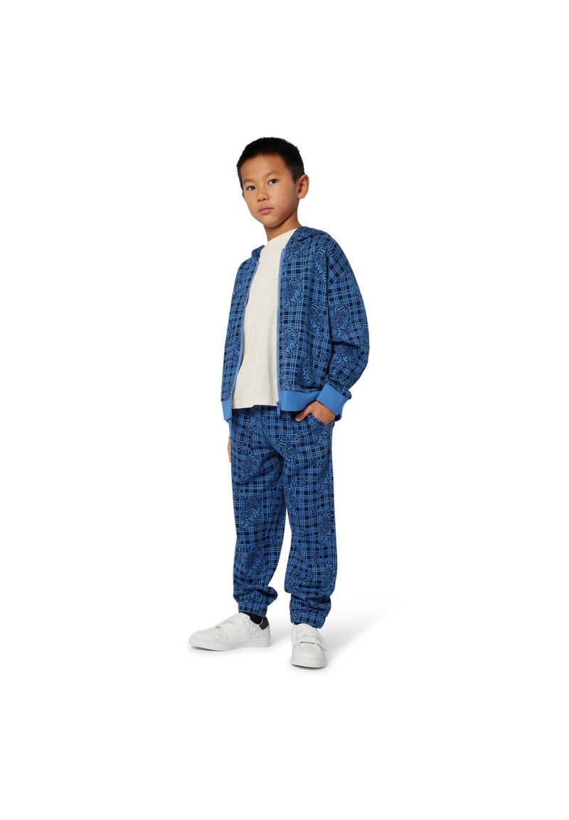 Ensemble de survêtement à motifs bleus avec une veste zippée et un pantalon à taille élastique, associé à des chaussures blanches. Texture lisse, design décontracté.