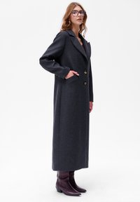 Lang, donkergrijze jas met een gekarteld kraag, twee gouden knopen, zijzakken en een gestructureerde stof. G gedragen met kniehoge bordeauxrode laarzen.