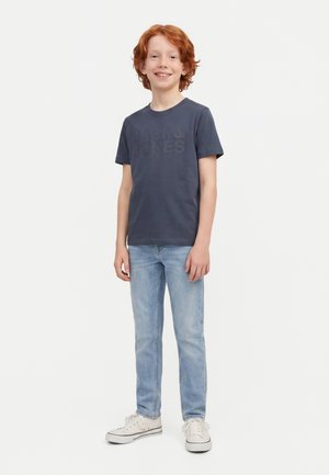Lächelnder rothaariger Junge steht, trägt ein dunkelblaues T-Shirt, hellblaue Jeans und weiße Sneakers vor einem einfarbigen weißen Hintergrund.