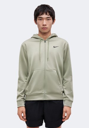 Giovane uomo che indossa una felpa con cappuccio verde chiaro con zip e piccolo logo nero Nike sul petto, e pantaloncini neri, in piedi davanti a uno sfondo semplice.