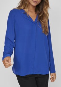 Blusa blu a maniche lunghe in tessuto leggero, con scollatura a V, pattina frontale e orlo leggermente arrotondato. Indossata con pantaloni scuri.