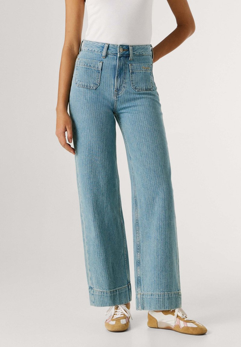 Femme portant un jean taille haute bleu clair à jambes larges avec poches plaquées devant et baskets beiges et blanches à lacets.
