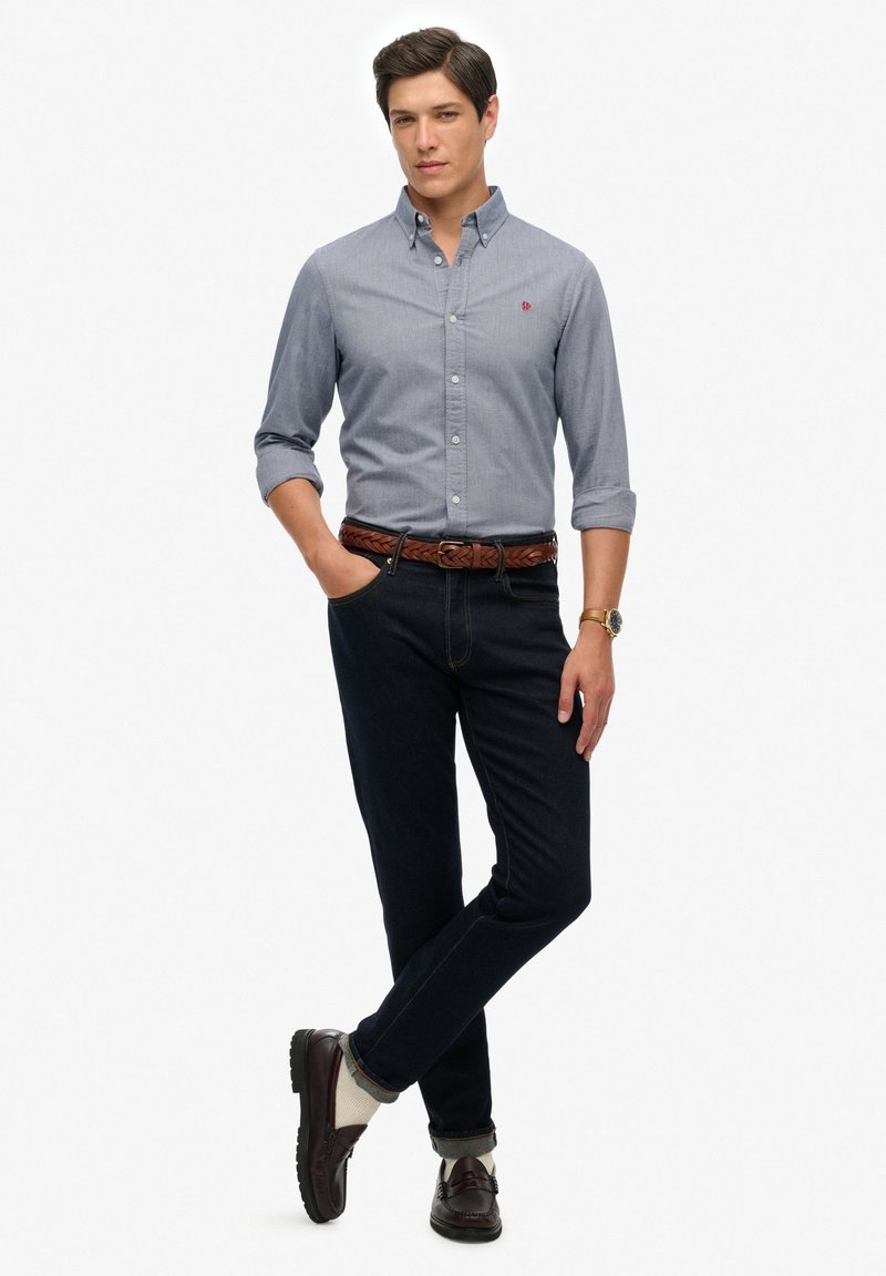Grijze overhemd met knopen en opgerolde mouwen, donkere jeans, bruine gevlochten riem en bruine loafers met lichte sokken. Eenvoudige, klassieke casual outfit.