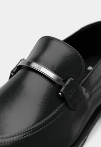 Sorte blanke skinnloafers med sølv metallrem som har gravert "BOSS"-logo på den øverste delen av skoen.