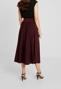 Burgundy pleated midi skirt made of soft fabric, featuring a high waistband and flowing design, paired with black strappy high-heeled shoes.