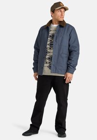 Men's blue jacket with a brown fur collar, two front pockets, worn over a patterned sweater and black pants, paired with a brown cap and black shoes.