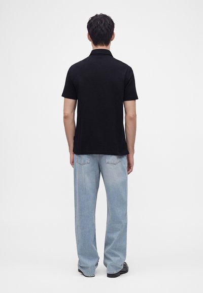 Homme debout, de dos, portant un polo noir à manches courtes, un jean bleu clair et des chaussures noires sur un fond blanc uni.