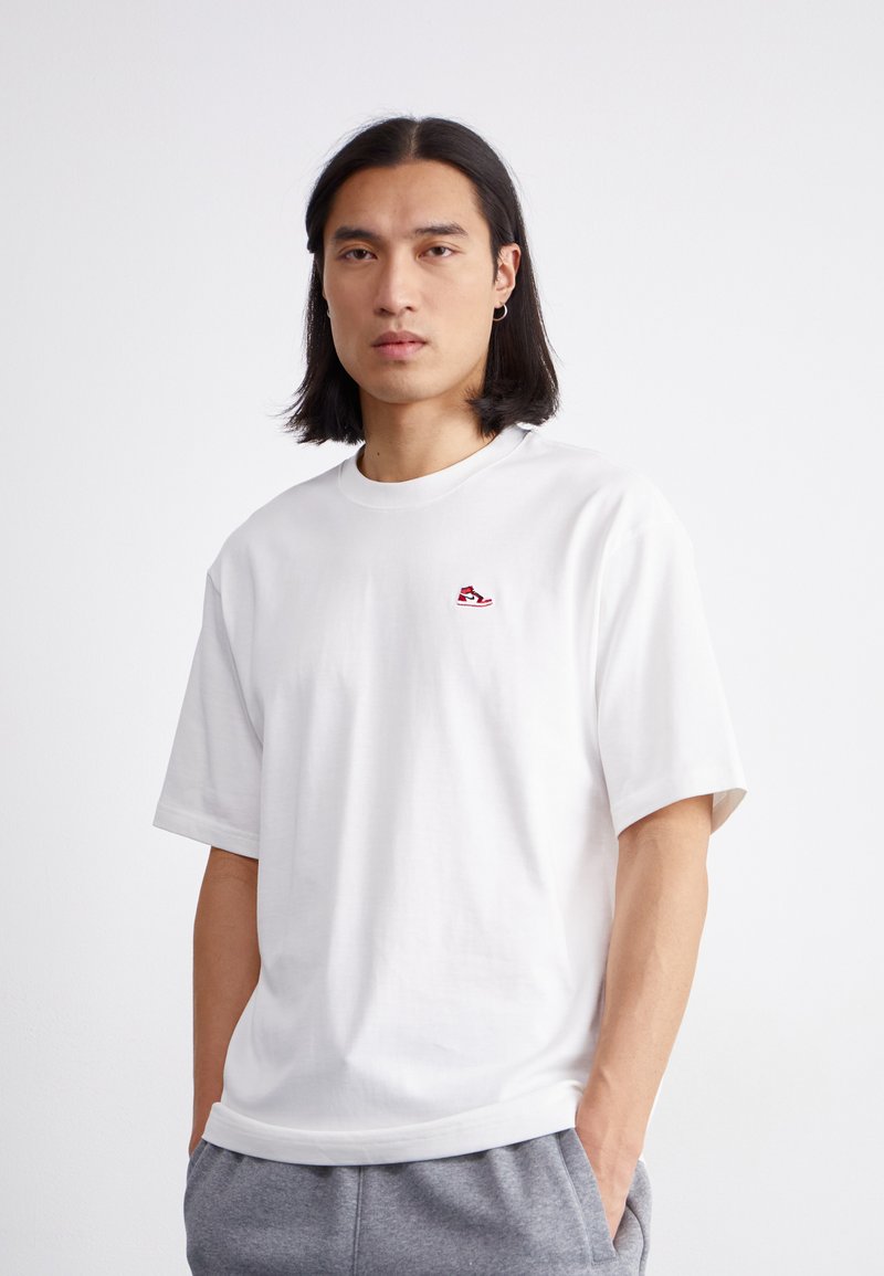 T-shirt bianco in cotone con maniche corte e scollatura rotonda, caratterizzato da un piccolo logo di sneaker rosso sul lato sinistro del petto.