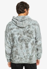 Sivá mikina s tie-dye vzorom a jemnou textúrou, s kapucňou, dlhými rukávmi a voľným strihom. Výrazné vyblednuté vzory po celom výrobku.