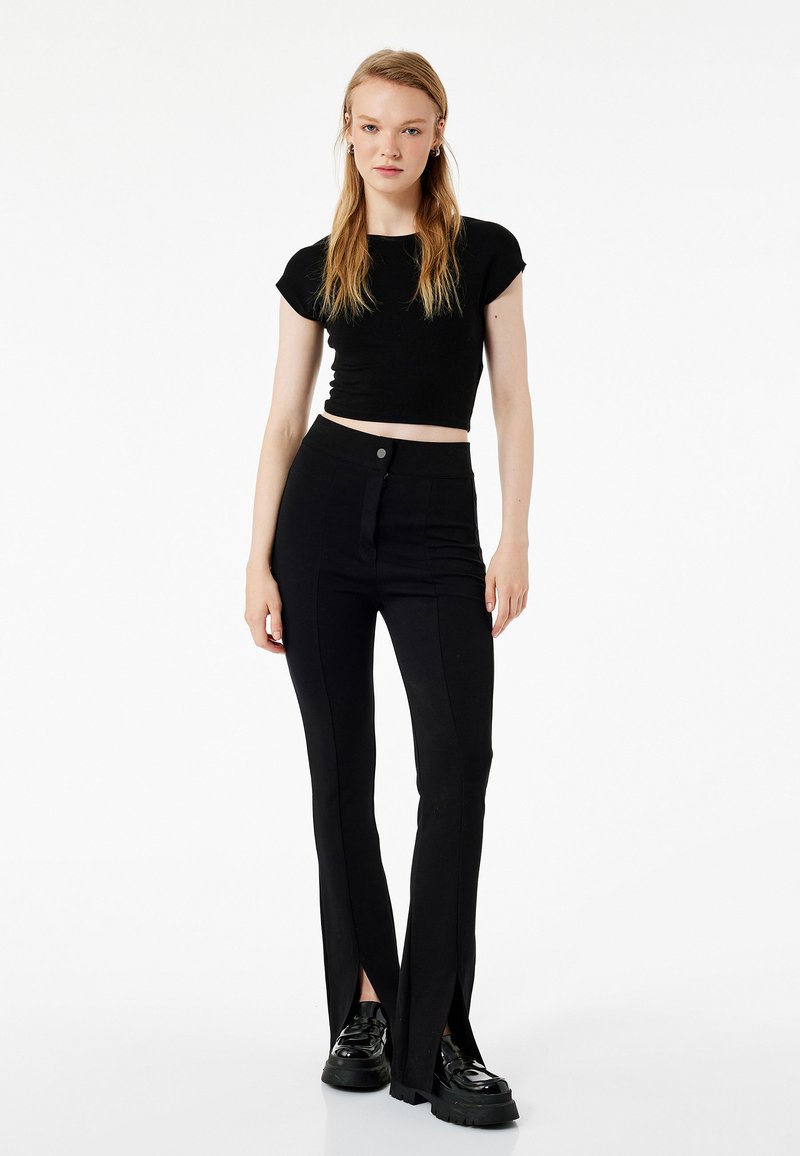 Schwarzes Crop-Top kombiniert mit hochgeschnittenen schwarzen Hosen mit seitlichen Schlitzen. Die Hosen haben ein elegantes Design und sind aus einem strukturierten Stoff gefertigt.