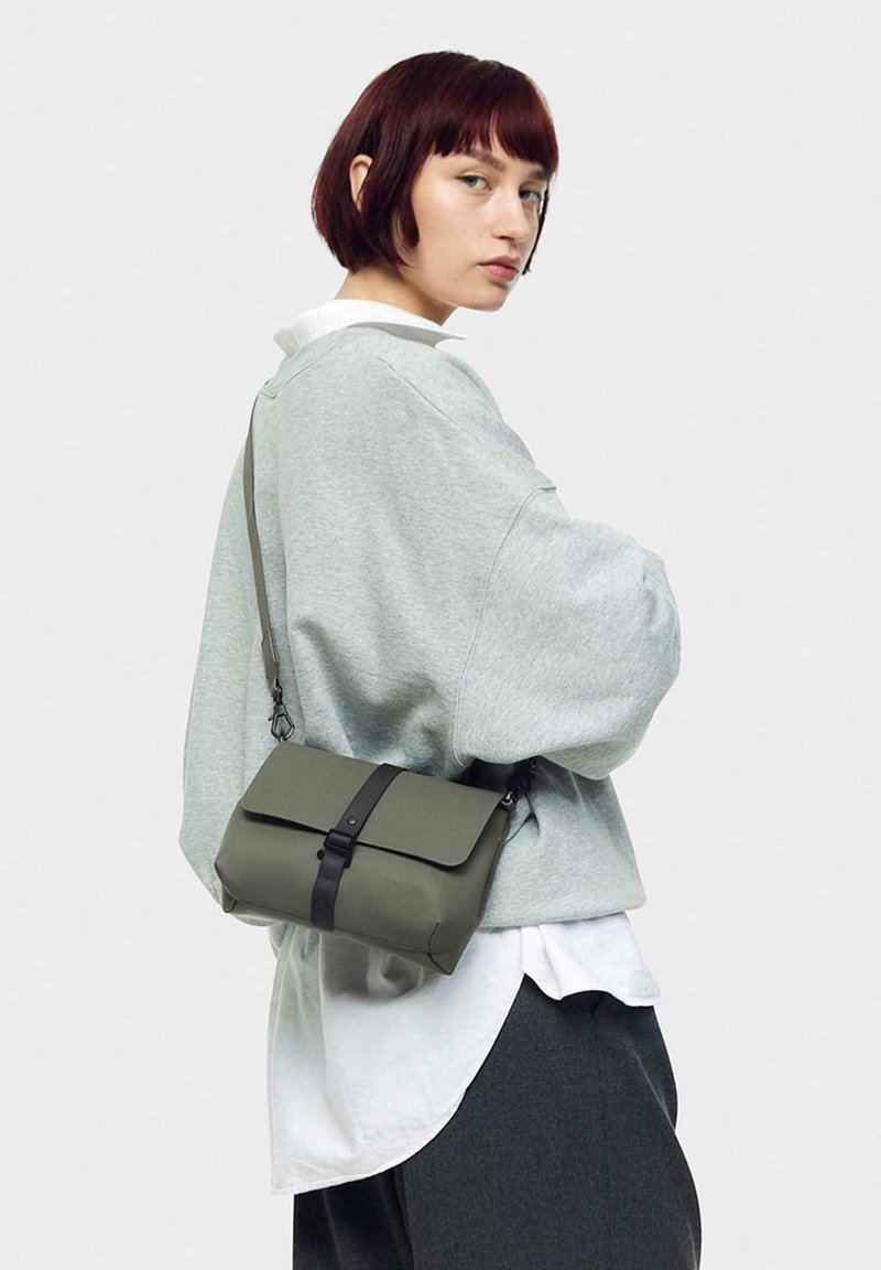 Grøn crossbody taske med en foldet klap, sort rem og spænde detaljer, parret med en lys grå oversized sweat-shirt og mørke bukser.