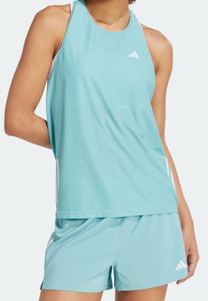 Kobieta ubrana w jasnoniebieski sportowy top bez rękawów oraz dopasowane szorty z białymi logo Adidas na jednolitym tle.