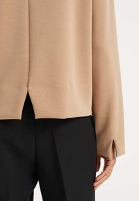 Top beige a maniche lunghe, tessuto morbido, spacchi laterali all'orlo, abbinato a pantaloni neri. Materiale liscio, design semplice.