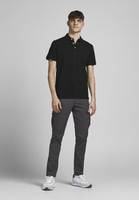 Zwarte polo met kraag en drieknoopssluiting, gecombineerd met grijze cargobroek met zijzakken. Witte sportieve schoenen.