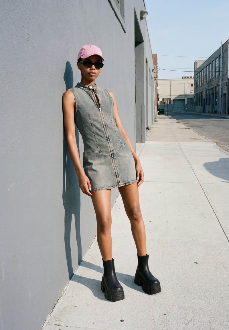 Mujer que lleva un vestido vaquero gris sin mangas, gorra rosa, gafas de sol negras y botas negras de plataforma apoyada en una pared gris en una acera.