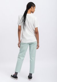 T-shirt blanche à manches courtes, associé à un pantalon vert clair ; le mannequin a de longs cheveux tressés et porte des baskets noires avec des chaussettes grises.