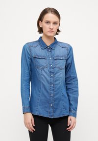 Πουκάμισο από τζην με κολάρο, μακριά μανίκια, δύο τσέπες στο στήθος και αντίθετο ραφί. Χρώμα: μεσαίο μπλε. Κουμπώνει μπροστά.