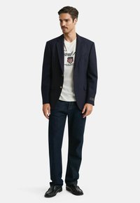 Marineblazer med guldknapper, båret over en hvid t-shirt med grafik, parret med mørke denimjeans og sorte loafers. Rene linjer og struktureret pasform.