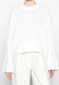 Weißer Strickpullover mit gerippter Struktur, weiten Ärmeln und kurzem Schnitt. Hergestellt aus leichtem Material und mit einem dezenten Muster versehen.