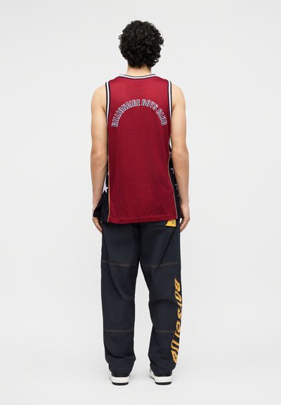 Άτομο με σγουρά μαλλιά που κοιτάζει προς τα πίσω, φοράει κόκκινη αμάνικη φανέλα "Billionaire Boys Club" και μαύρο παντελόνι με κίτρινο κείμενο στο πόδι.