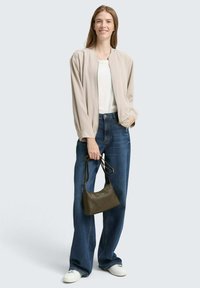 Hellbeige Bomberjacke über einem weißen Oberteil, kombiniert mit blauen, weit geschnittenen Jeans und weißen Sneakers. Hält eine kleine olivgrüne Handtasche.