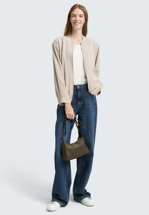 Hellbeige Bomberjacke über einem weißen Oberteil, kombiniert mit blauen, weit geschnittenen Jeans und weißen Sneakers. Hält eine kleine olivgrüne Handtasche.