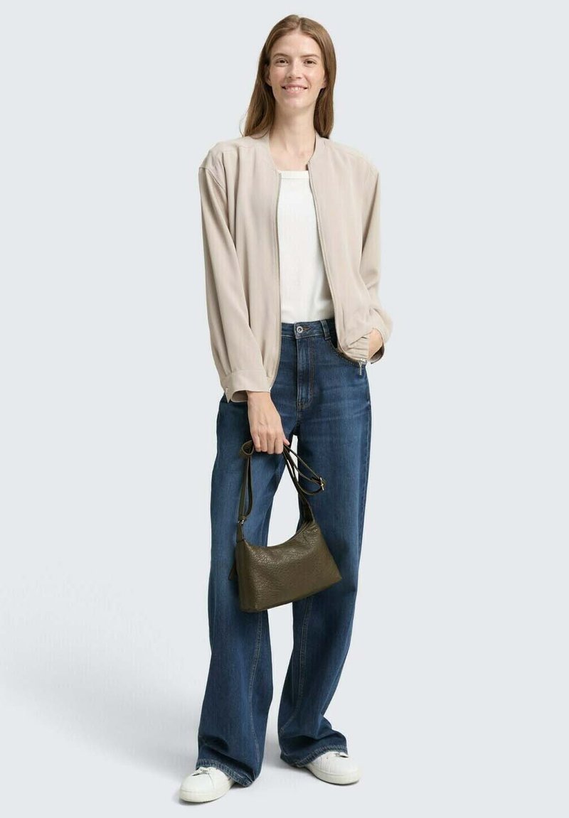 Hellbeige Bomberjacke über einem weißen Oberteil, kombiniert mit blauen, weit geschnittenen Jeans und weißen Sneakers. Hält eine kleine olivgrüne Handtasche.
