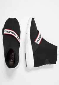 Black knit slip-on sneakers with a white midsole, featuring a red and white branded strap across the upper and a textured knit finish.