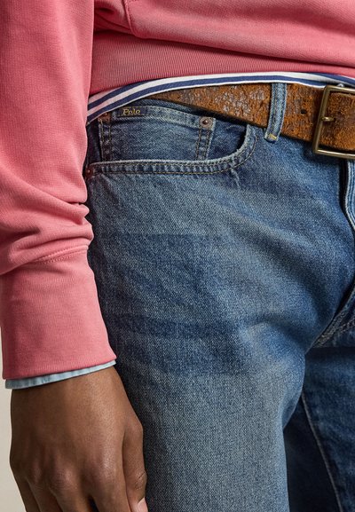 Közeli kép egy személyről, aki kék farmernadrágot visel barna bőr övvel és rózsaszín hosszú ujjú inggel, amelynek kék mandzsettája látható.