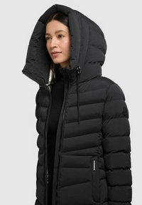 Giubbotto puffer nero con un ampio cappuccio strutturato, design trapuntato, zip frontale e dettaglio con logo sulla manica. Texture liscia.