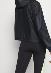 Black, hooded jacket with a cropped design and smooth, shiny texture. It features elastic cuffs and a relaxed fit, paired with black leggings.