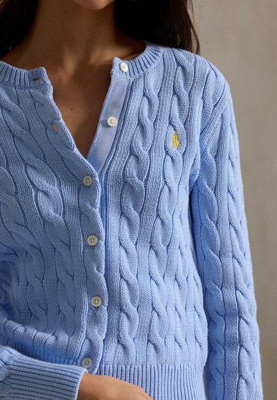 Nő világoskék, kötött mintázatú kardigánban, fehér gombokkal és a mellkasán kis sárga hímzett logóval.