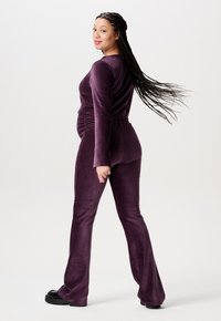 Lilla, udvidet jumpsuit lavet af blødt stof, med lange ærmer og rynkede detaljer ved taljen, båret af en person med fletninger.