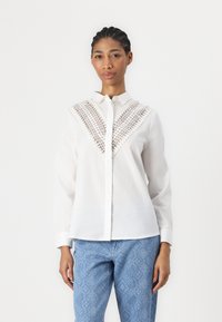 Camicia bianca con bottoni, caratterizzata da un design decorativo all'uncinetto sul petto, con maniche lunghe e vestibilità comoda. Abbinata a jeans azzurri con motivo.
