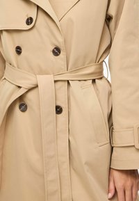 Trench-coat beige avec un design croisé, boutons marron, taille ceinturée et silhouette épurée. Tissu doux et poches latérales.