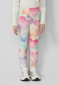 Kind trägt weiße Turnschuhe und Leggings mit bunten Aquarellherzen, steht vor einem einfarbigen hellgrauen Hintergrund.