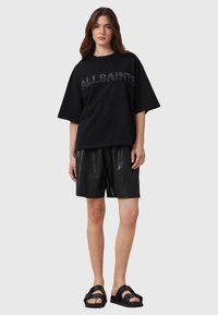 Camiseta negra de gran tamaño con "ALLSAINTS" en letras texturizadas, combinada con pantalones cortos de cuero negros y sandalias negras con correas gruesas.