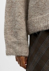 Pull en laine beige avec un tricot texturé. L'ourlet et les poignets côtelés offrent un ajustement parfait. Main reposant sur un tissu à carreaux brun.
